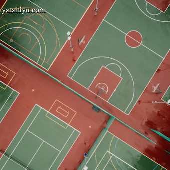 Aerial view of outdoor sports courts displaying multiple basketball and tennis courts positioned side by side. The courts are painted with green playing surfaces and red surrounding areas, with white and yellow boundary lines. The basketball courts feature hoops and painted three-point arcs and key areas, while the tennis courts include service lines and baselines.