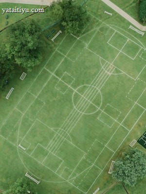 An aerial view of a sports complex featuring multiple sports fields drawn on green grass. The layout includes soccer and tennis courts, with pathways surrounding the area. Trees and surrounding buildings are visible, along with a swimming pool section on the left side.