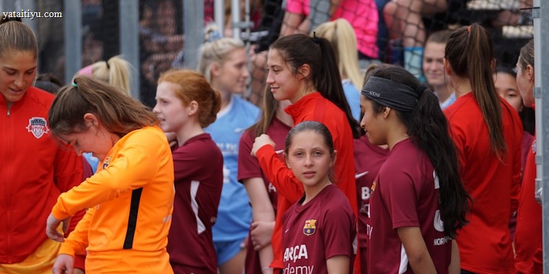 A group of young female athletes in sports attire are gathered together, some wearing red jackets or maroon jerseys with the logo of a soccer academy. They appear to be standing in front of a wire fence with spectators in the background, dressed in casual clothing.