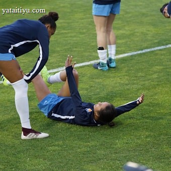 A group of athletes in sportswear are on a green soccer field. Two individuals are interacting closely; one helps the other who is lying on the grass. Nearby, other athletes are stretching.
