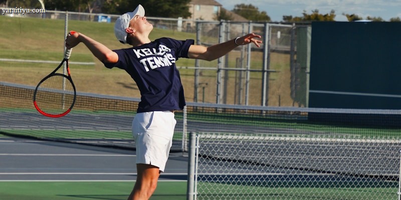 A tennis player is captured mid-air while serving at an outdoor tennis court. The player is wearing a navy blue shirt with 'Keller Tennis' printed on it, white shorts, and bright green sports shoes. A tennis racket is in the player's right hand, while the tennis ball is in the air above. The background includes a chain-link fence, a field, trees, and a partly cloudy sky.