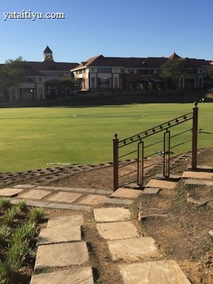 A well-maintained sports field is bordered by a stone pathway and decorative railing in the foreground, with large buildings in the background. A water sprinkler is active on the field, and two small soccer goals are visible at the far end. The scene is illuminated by bright sunlight, casting clear shadows.