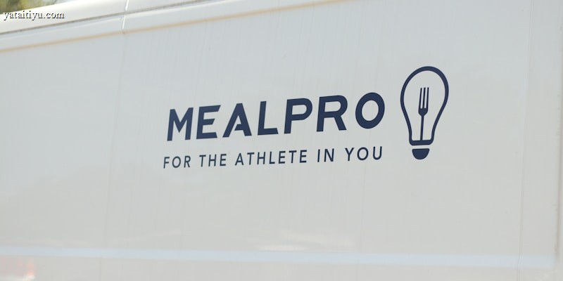 The side of a white vehicle displays the meal service logo 'MEALPRO' with the tagline 'For the Athlete in You'. Next to the text is a graphic combining a lightbulb and a fork.